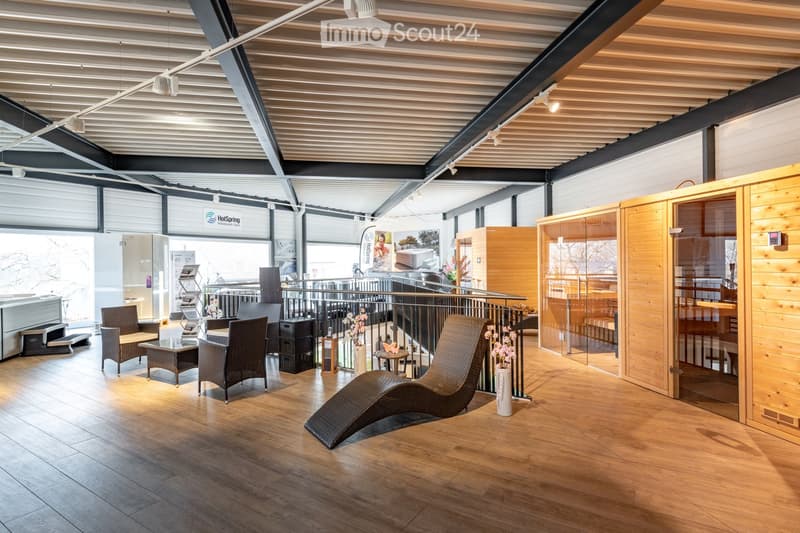 Modern, spacious room with wooden flooring, large windows, and a sauna area. Features include several wicker chairs, a glass coffee table, a vase with flowers, and a display screen.
