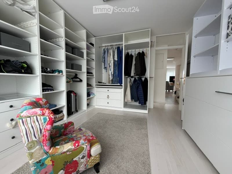 A well-organized walk-in closet with white built-in wardrobes, shelves, and drawers. A colorful armchair is placed on a light-colored carpet, and the space features wooden flooring and ample storage.