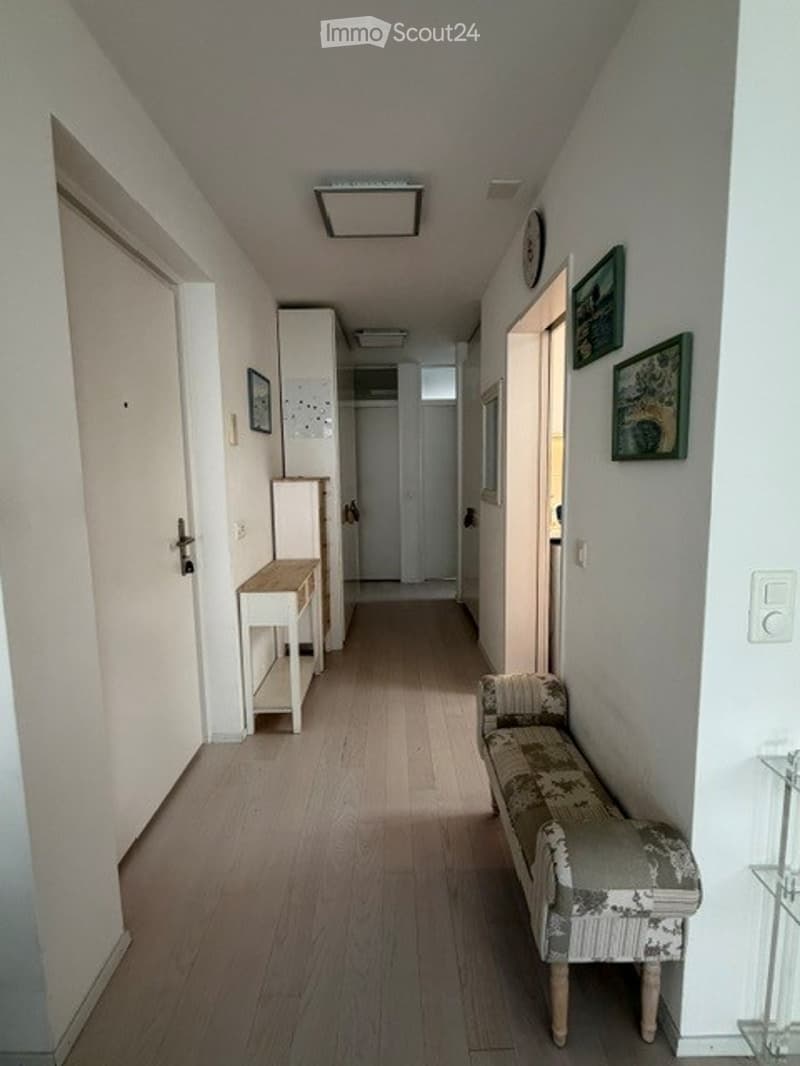 A narrow hallway with light wooden flooring, white walls, and a few pieces of furniture including a small bench and a small table. The hallway leads to other rooms and has framed pictures on the walls.