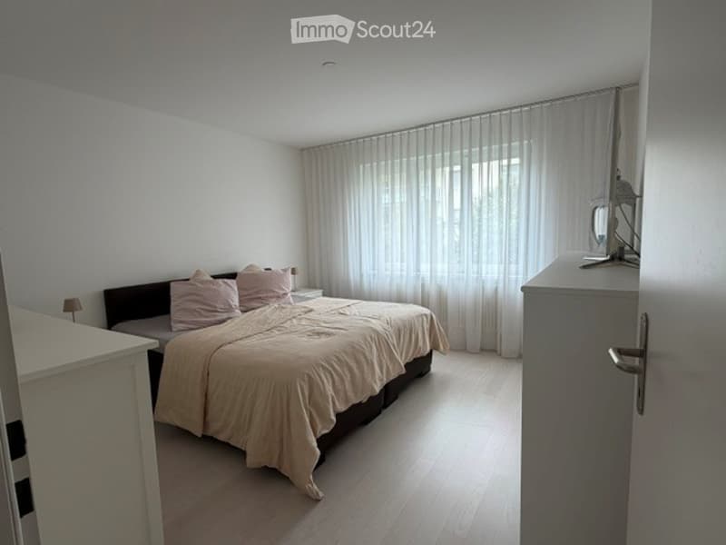 A modern bedroom with a large bed, beige bedding, pink pillows, a nightstand with a lamp, a mirror on a cabinet, and sheer white curtains covering a window.