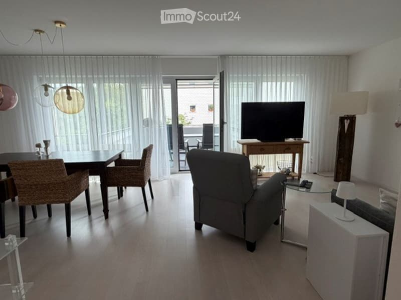 Bright living and dining area with large windows and sliding doors leading to a balcony. Features a grey sofa, dining table with four wicker chairs, a television on a wooden stand, and modern lighting.