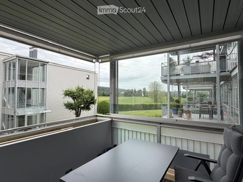 Modern balcony with a sleek black table and a comfortable chair, offering a scenic view of a green landscape and neighboring buildings.