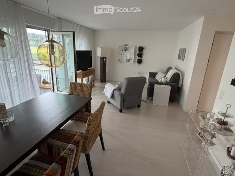 Open-plan living and dining area with modern furniture, large windows, and a balcony view.