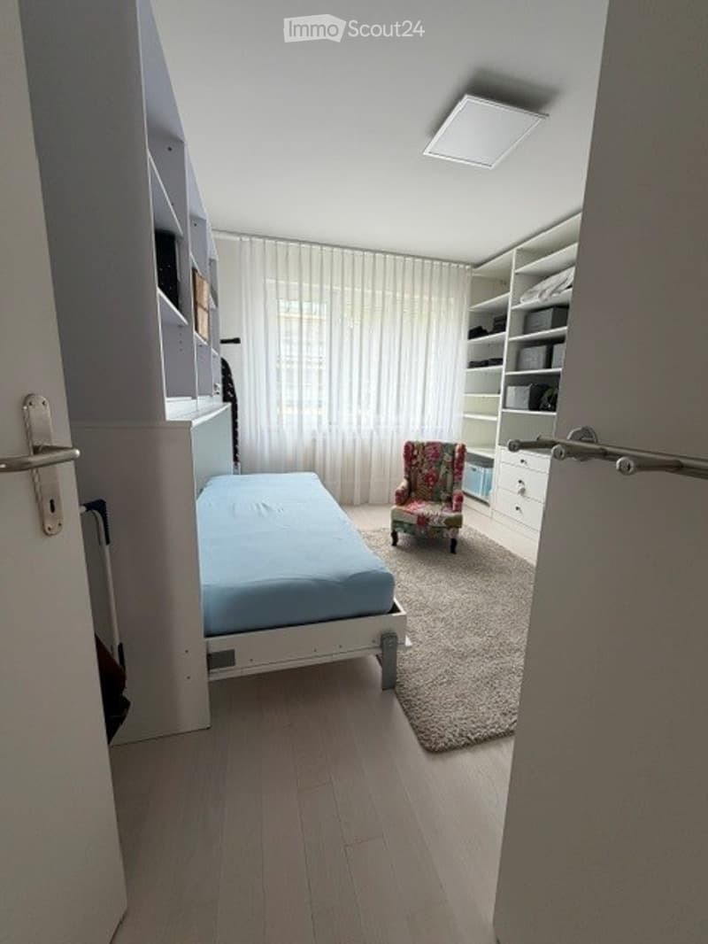 A small, modern bedroom with a single bed, a colorful armchair, built-in wardrobe, shelves, and a large window with white curtains.