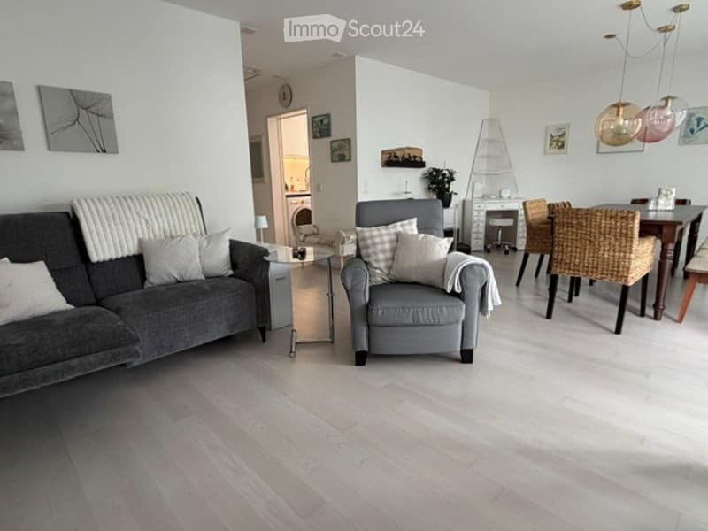 Modern living room with light wood flooring, grey sofa, armchair, coffee table, dining area with chairs, and decorative wall art.