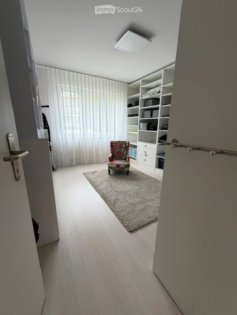 A small, well-lit room with light wooden flooring, a patterned armchair on a beige rug, built-in white shelving units, and sheer white curtains covering a large window.