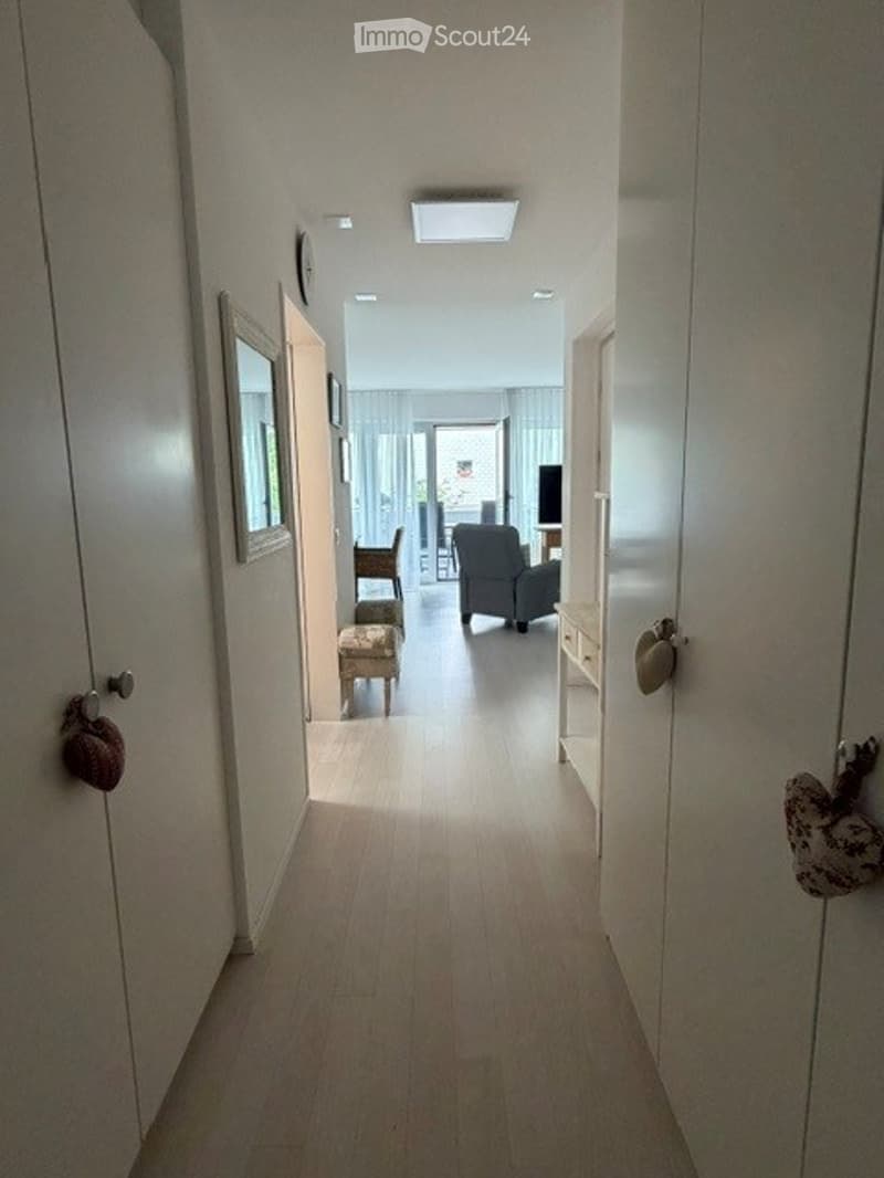 A well-lit hallway with wooden flooring, white walls, and several doors. A small seating area is visible at the end of the hallway, featuring a sofa, chairs, and a coffee table.