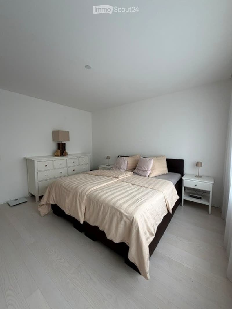Modern bedroom with a large bed, nightstands, lamps, and a dresser. Light-colored bedding and white walls.