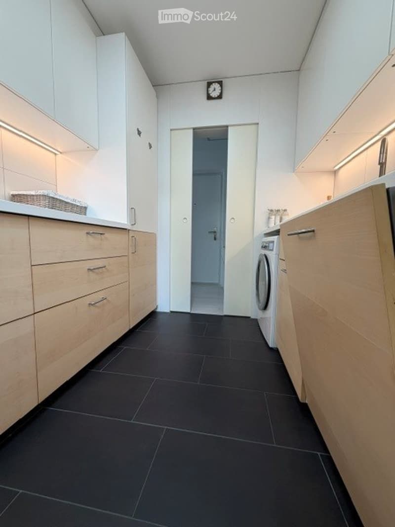 Modern kitchen with light wooden cabinets, black tiled floor, and a private washing machine visible in the background.