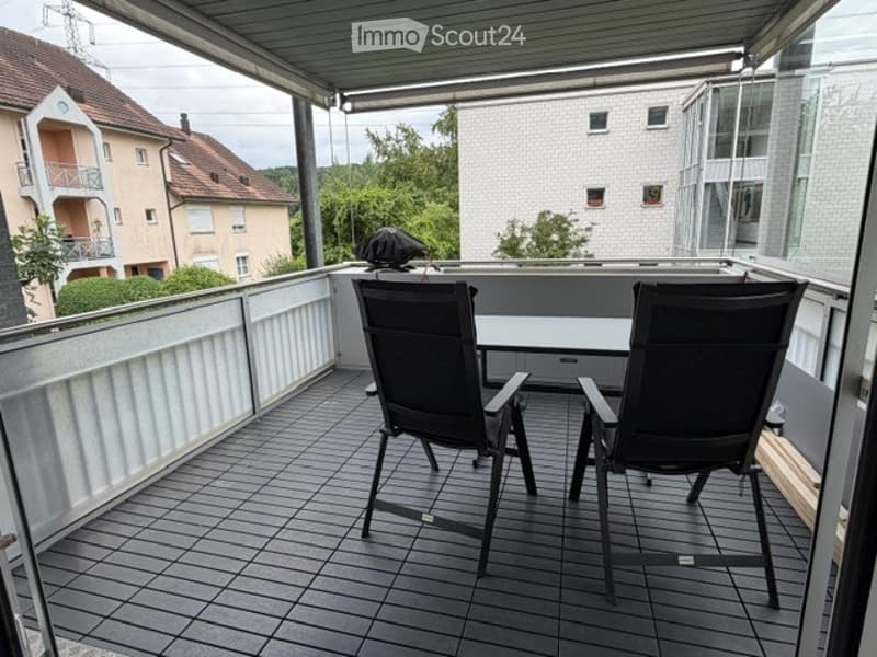Balcony with a covered roof, tiled floor, two black folding chairs, and a small table. The view shows neighboring buildings and greenery.