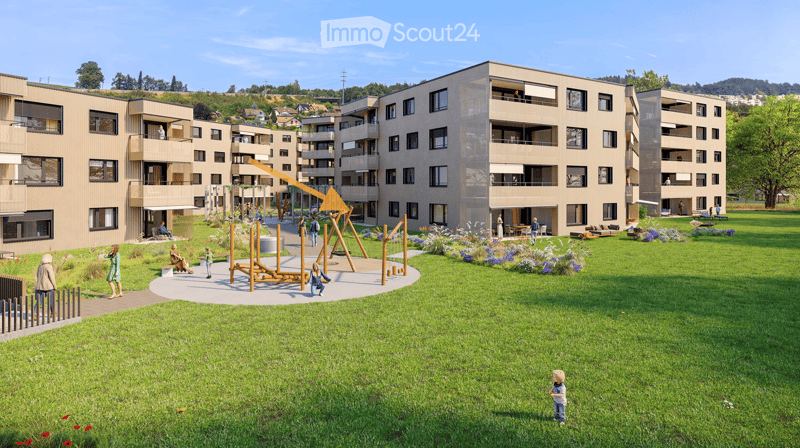 A modern residential complex with a spacious green lawn, playground equipment, and several people enjoying the area. The buildings have contemporary architecture with balconies and large windows.