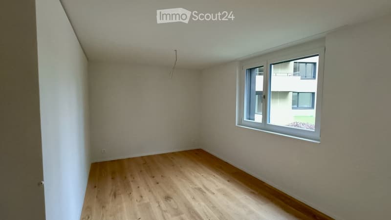 Empty room with wooden flooring, white walls, and a large window with a view of a neighboring building.