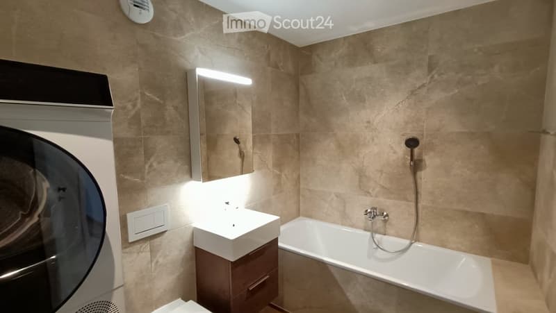 Modern bathroom with beige marble tiles, a white sink with a dark wooden cabinet, a bathtub with a handheld shower, and a large mirror with built-in lighting.