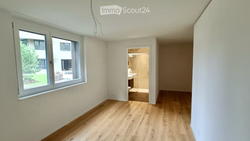 Empty room with wooden flooring, white walls, and large windows. A doorway leads to a small bathroom with a shower and sink.