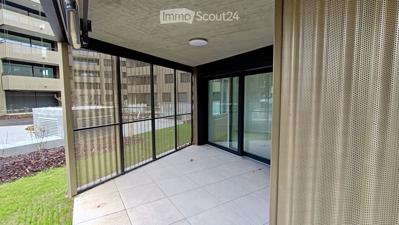 Modern balcony with large glass panels, tiled floor, and a view of a contemporary building and landscaped area.