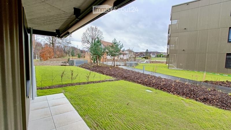 Well-maintained garden area with neatly trimmed grass, mulch beds, small trees, and a paved pathway leading to a modern building.