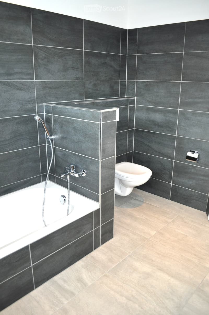 Modern bathroom with dark grey tiled walls, a white bathtub with a glass shower enclosure, a toilet, and a chrome faucet.