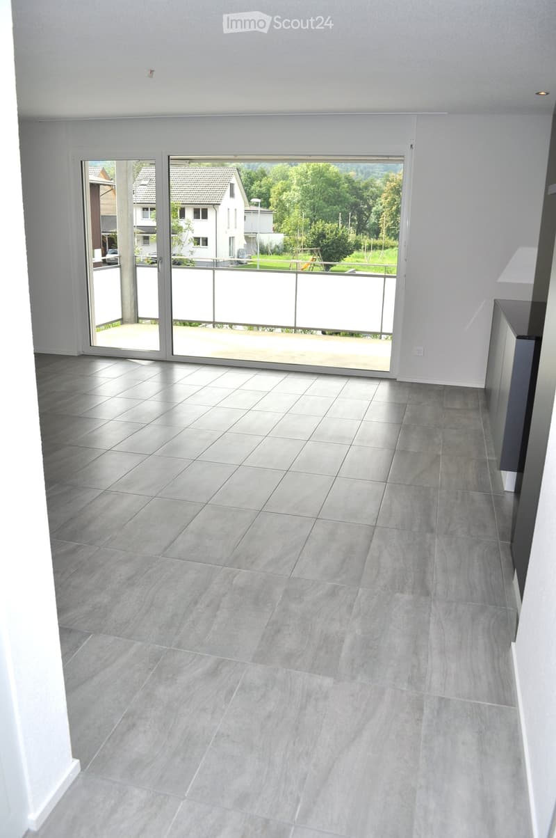 Empty room with grey tiled floor and large sliding glass doors leading to a balcony with a view of the neighborhood.