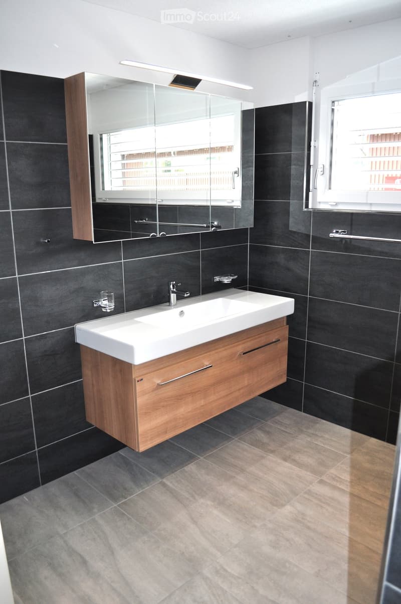 Modern bathroom with dark tiled walls, a white sink with wooden cabinet, a large mirror, and a window providing natural light.