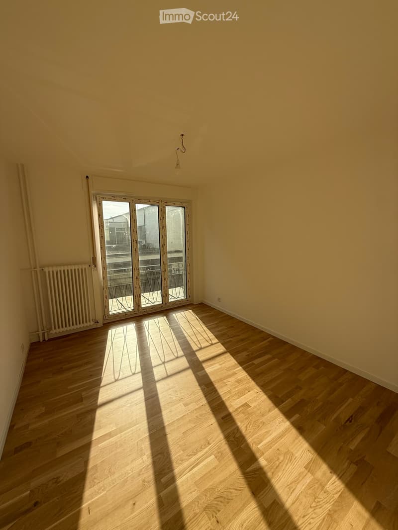 Empty room with light wooden flooring and large glass sliding doors leading to a balcony. Light yellow walls and a radiator visible on the left side.