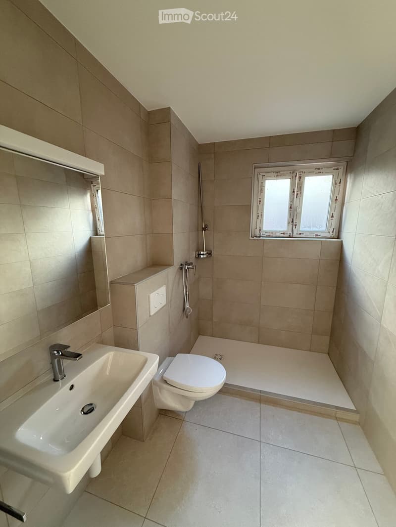 Modern bathroom with beige tiles, a white sink, toilet, and a shower area with a glass window above.