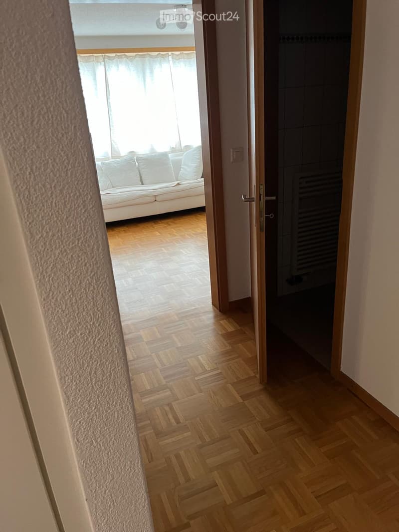A hallway with wooden floors, open doors leading to rooms, white walls, a sofa with white pillows in the living room, and a window with white curtains.