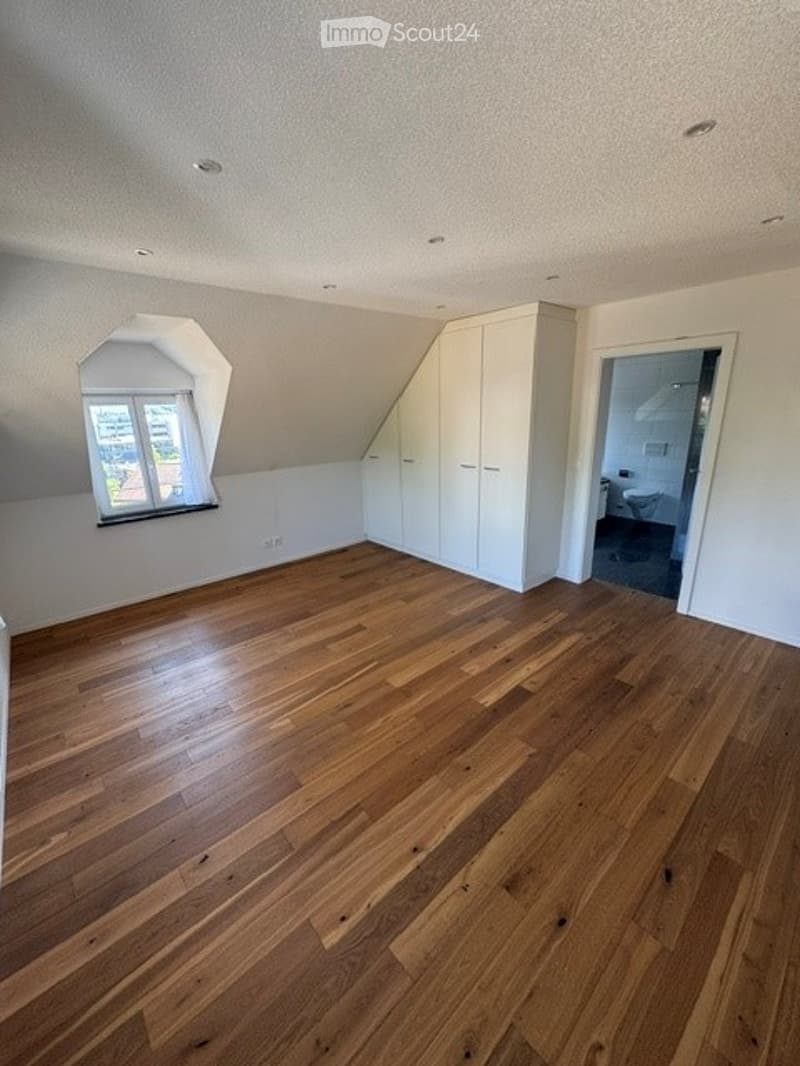 Empty attic room with wooden flooring, white walls, built-in closet, and a window with a view of the outside.