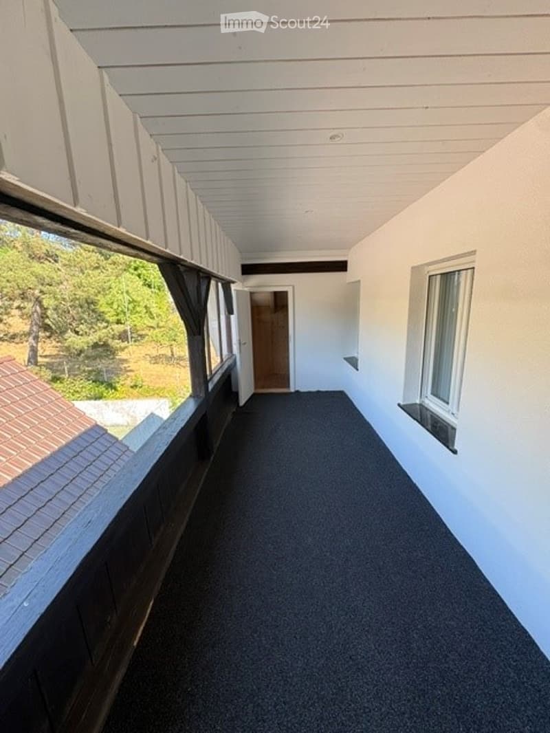 Empty hallway with white walls, carpeted floor, and large windows offering a view of the outdoors.