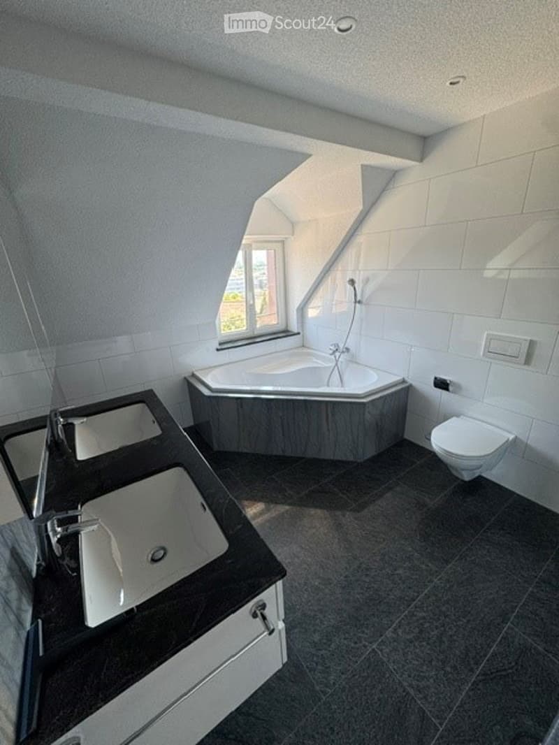 Modern bathroom with a corner bathtub, double sink vanity, and toilet. The space features dark tiled floors and white tiled walls, with a skylight window providing natural light.