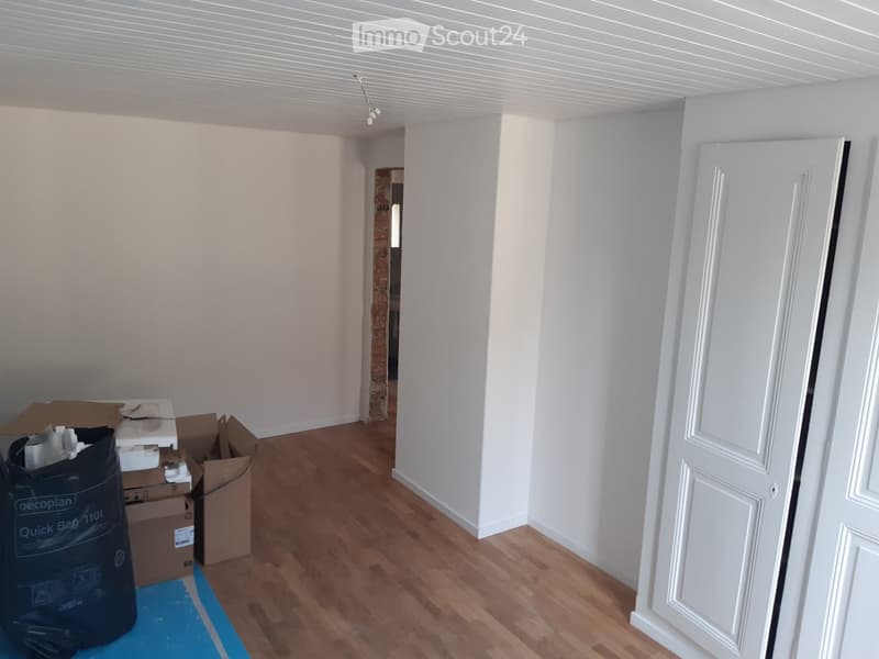Empty room with white walls, wooden flooring, and a white door. Cardboard boxes and a black bag are visible in the corner.