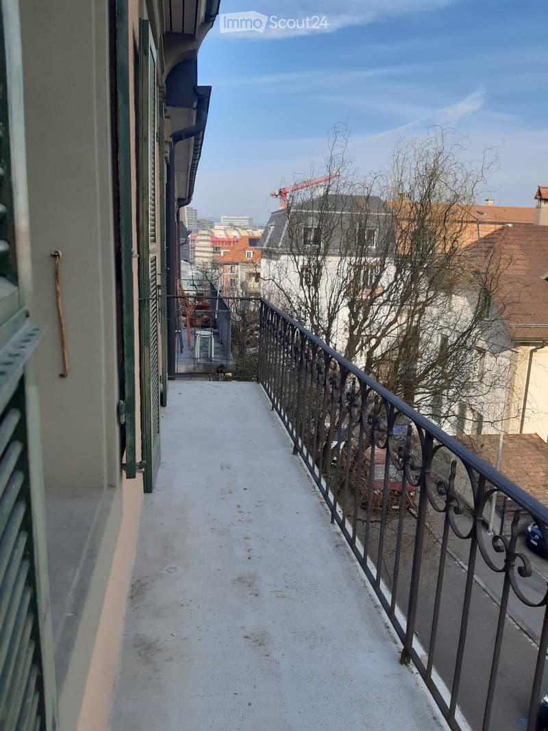 Balcony with metal railing, concrete floor, overlooking cityscape with buildings and trees.