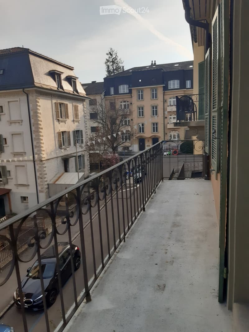 Balcony with metal railing overlooking a narrow street lined with multi-story residential buildings, some with balconies and greenery.