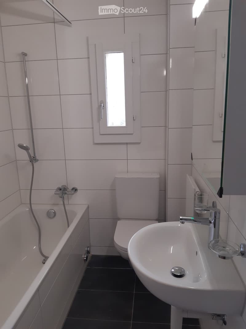 Modern bathroom with white tiles, bathtub, toilet, sink, and a small window above the toilet.