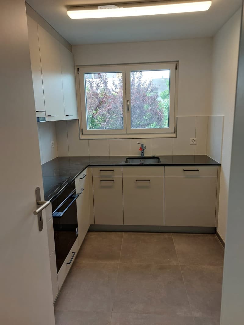 Empty kitchen with white cabinets, black countertops, a sink, and a window overlooking greenery.