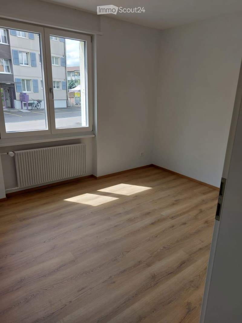 Empty room with wooden flooring, large window with blinds, radiator, and visible sunlight streaming in.