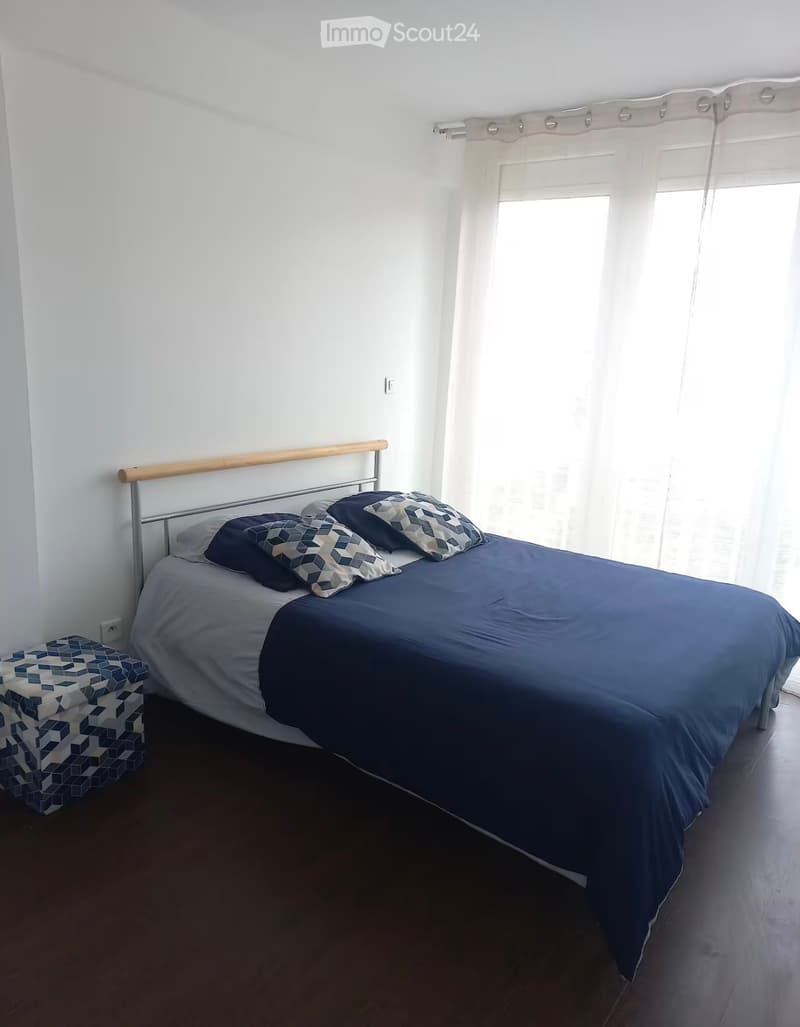 A modern bedroom with a double bed, dark blue bedding, patterned pillows, and a matching stool. The room features white walls, wooden flooring, and sheer white curtains on a large window.