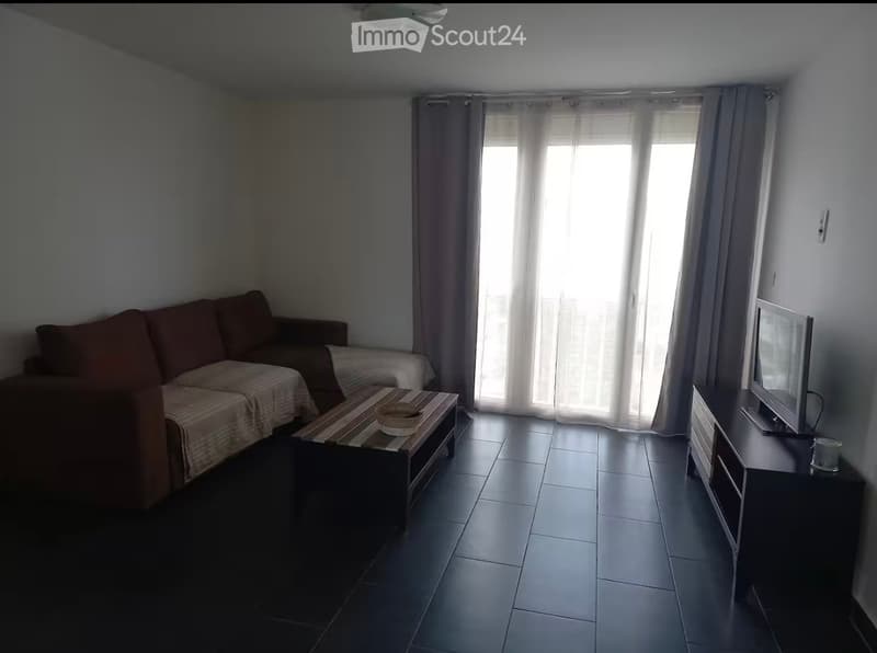 Living room with a sofa, coffee table, TV stand, and a small table with a bowl on it. The room has large windows with curtains and tiled flooring.