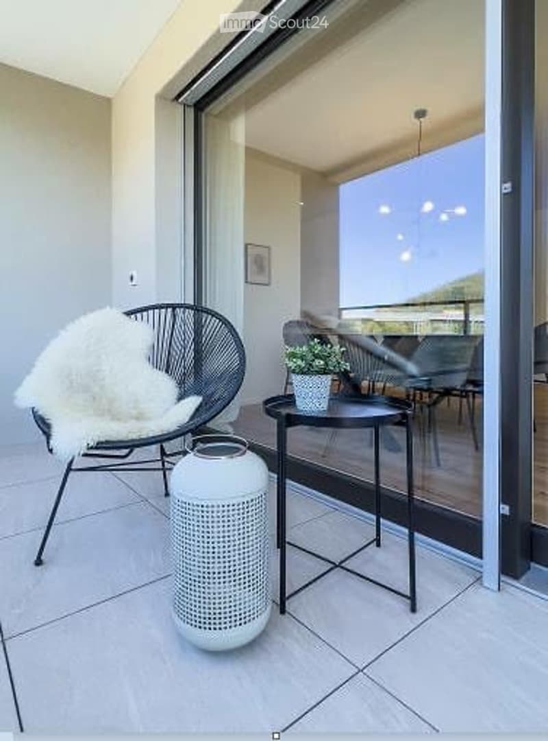 Modern balcony with large glass sliding doors, white tiled floor, black round table, white chair with fur throw, small potted plant, and white decorative vase.