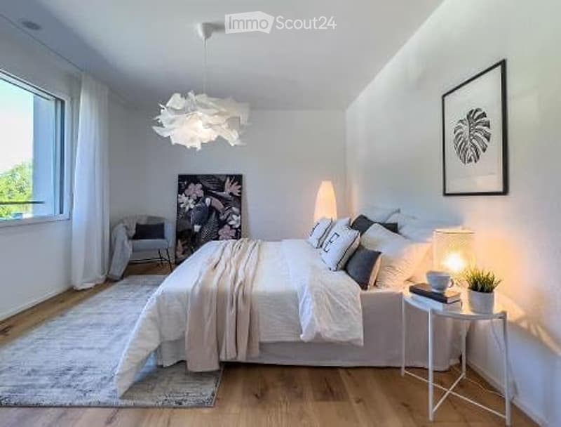 Modern, minimalist bedroom with a large bed, nightstand, chair, and decorative elements including a framed picture and a potted plant. The room features white walls, wooden flooring, and large windows with sheer curtains.