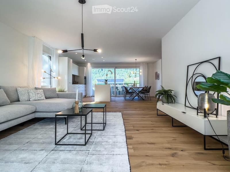 Modern living room with a grey sectional sofa, black coffee tables, a dining area with chairs and a table, large windows, and decorative plants.