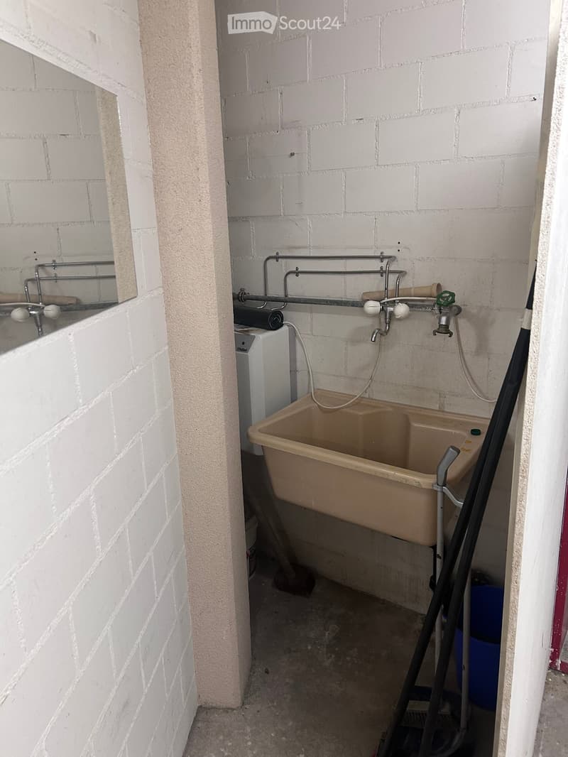 A small bathroom with a sink, faucet, pipes, and a washing machine. The walls are made of white bricks, and the floor is concrete.