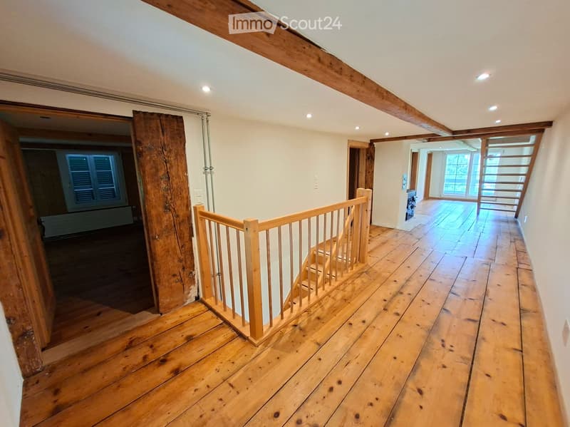 Wooden staircase with a wooden railing, wooden floors, and exposed wooden beams on the ceiling. The hallway leads to another room with large windows and a tiled floor.