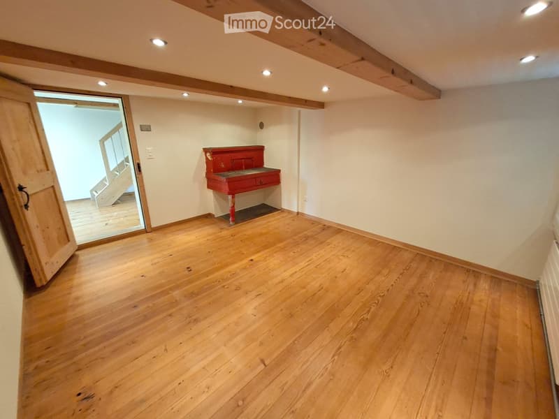 Empty room with wooden flooring, white walls, exposed wooden beams on the ceiling, and a small red console table against one wall. An open doorway leads to another room with wooden flooring and a staircase.