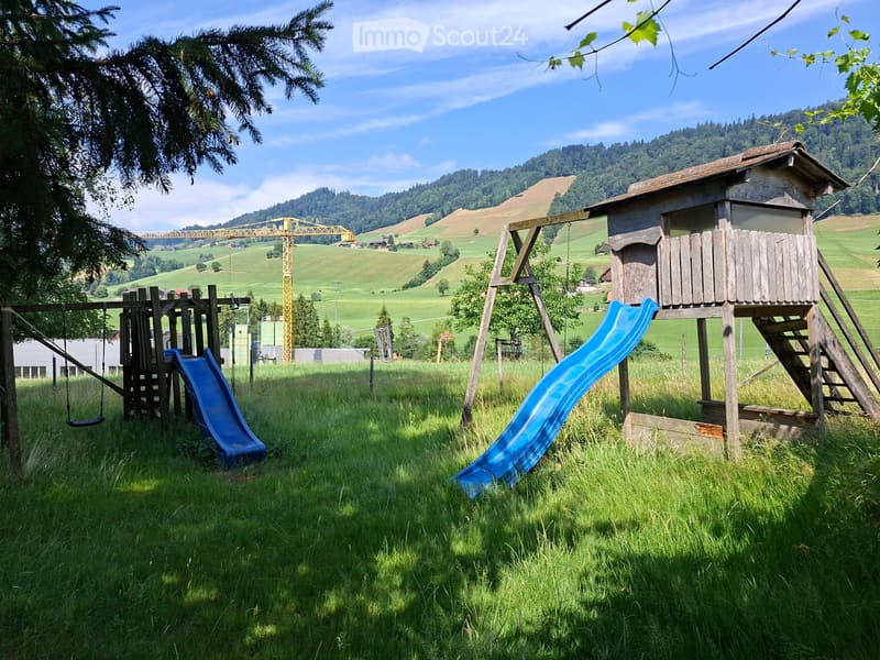 A grassy outdoor area with a wooden play structure featuring a swing and a blue slide, surrounded by trees and hills.