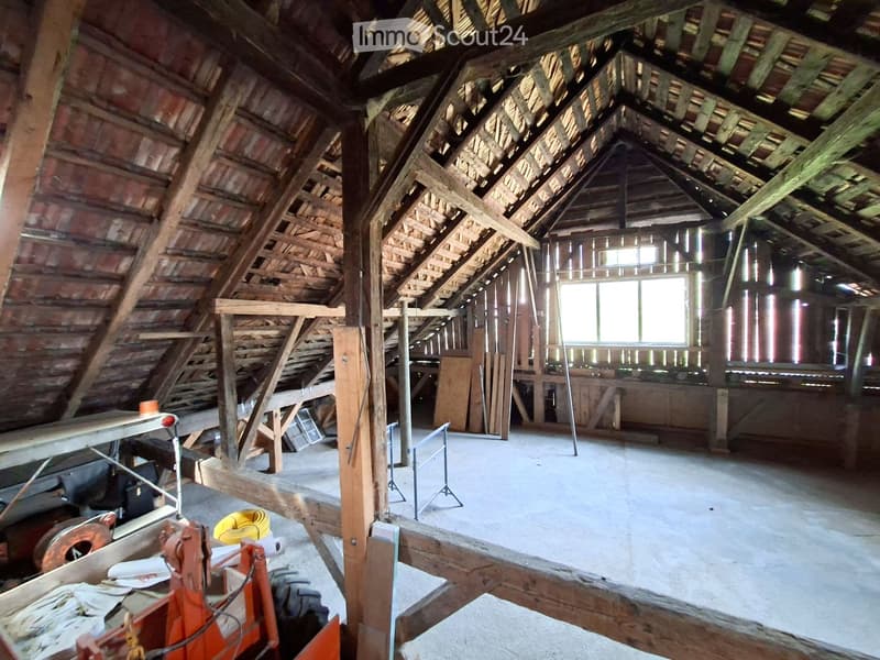 An attic space with exposed wooden beams and rafters, unfinished walls, and a concrete floor. There are some construction materials and tools scattered around, including a workbench, a ladder, and a window letting in natural light.