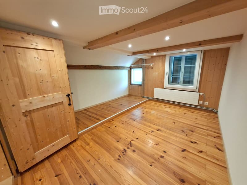Empty room with wooden flooring, exposed wooden beams on the ceiling, and two windows with shutters. The room has a large wooden door on the left side and a radiator on the right wall.