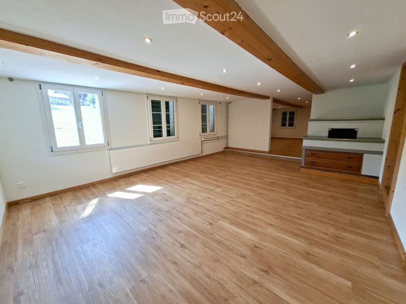 Empty room with wooden flooring, white walls, exposed wooden beams on the ceiling, and large windows allowing natural light to enter.