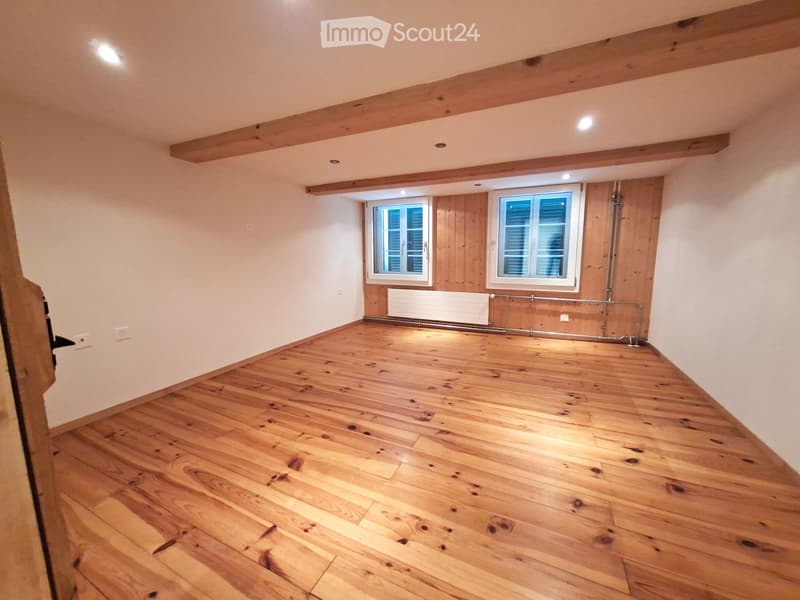 Empty room with wooden flooring, white walls, exposed wooden beams on the ceiling, and two windows with shutters.