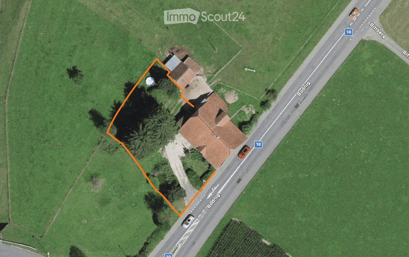 Aerial view of a two-story house with a red-tiled roof, surrounded by greenery and located near a road.