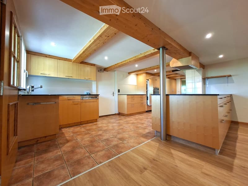 Modern kitchen with light wooden cabinets, stainless steel appliances, and a central island. The space features a mix of tiled and wooden flooring, with exposed wooden beams on the ceiling.